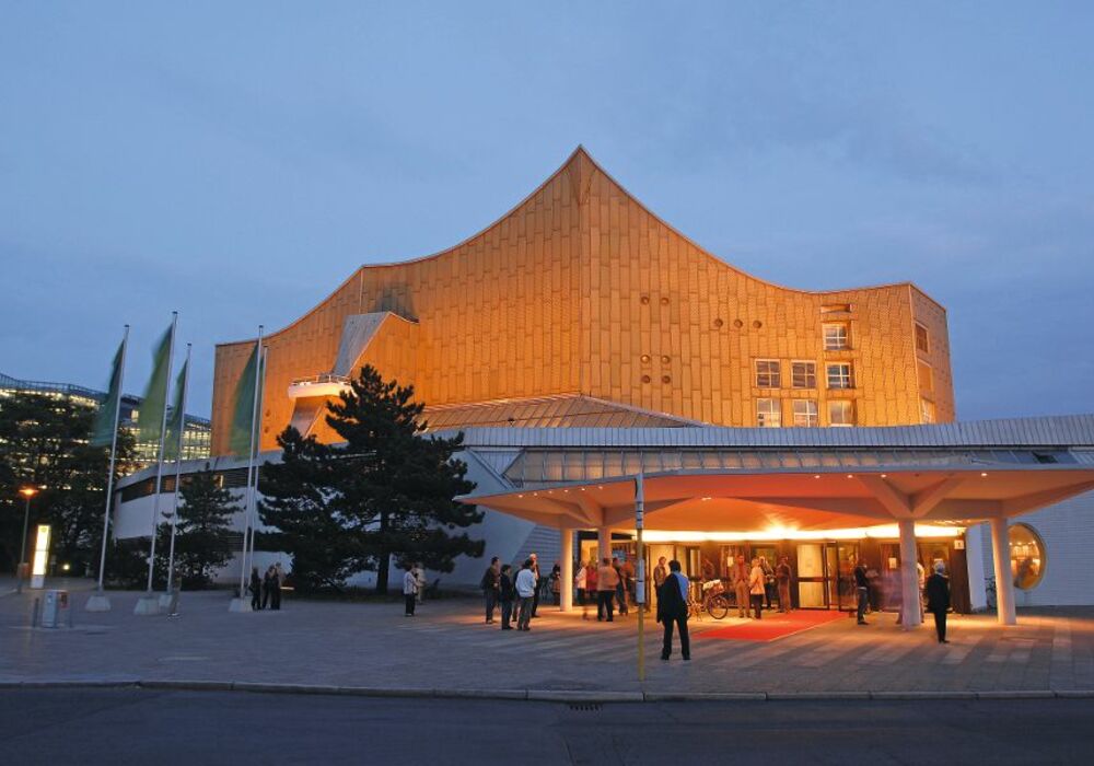 Berliner Philharmonie bei Abenddämmerung, beleuchtet, mit Besuchern am Eingang.